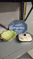 Three ceramic dishes on a metal shelf: blue and white platter, green leaf-shaped dish, cream square lidded dish with black script