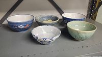 Five ceramic soup bowls in a row showing variety of designs and colors, on a metal shelf.
