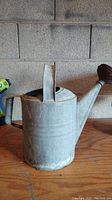 Side view of antique metal watering can showing spout and handle with rust and wear.