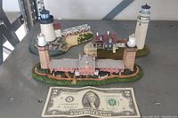 Three Cape Cod lighthouse themed miniature landscape models displayed on a table beside a US two dollar bill for scale.