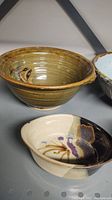 Three pottery items: 10-inch mustard-brown bowl, 7-inch cream and purple glazed shallow bowl, and partially visible 9-inch pale blue bowl