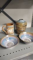Four handmade pottery items: two spiral pattern bowls, a decorated mug with handle, and a rounded vase with horizontal glaze.
