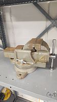 Front side view of the heavy-duty bench vise placed on a metal shelf, showing the jaws, handle, and base