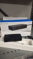 Microsoft Surface Dock in front of original box showing branding and product image, power cable attached.