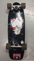Top view of mini longboard skateboard showing black deck with white spade graphic and Punked logo on the tail, brown wheels and metal trucks.
