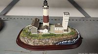 Overall view of Danbury Mint Montauk Island Lighthouse miniature showing the lighthouse tower, surrounding buildings, and rocky base with greenery.