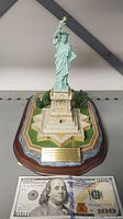 Full view of the Statue of Liberty miniature model on wooden base, next to a $100 bill for size reference