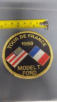 Full view of round embroidered patch with tape measure showing near 3 inches diameter. Black background with gold embroidered 'Tour De France 1999', crossed American and French flags, and 'Model T Ford' text.