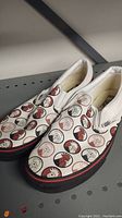Pair of Vans slip-on shoes showing rooster head pattern on canvas, red and black sole.