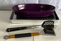 Four barbequing and baking accessories including a purple oval roasting pan, rectangular tray with grill rack, slotted spatula, and dual-sided barbecue brush.