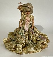 Full view of antique doll in flowing yellow satin and floral dress with ruffles and bonnet.
