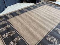 View of full rug showing beige textured center with black border featuring repeated pineapple motifs in beige.