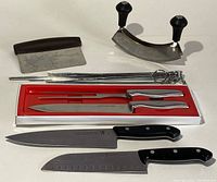 Photo showing full set of knives, mezzaluna chopper, dough scraper, metal skewers, and small forks against white background.