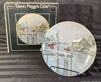 Collector plate in front of original display box with artwork and title visible, shown on black fabric background.