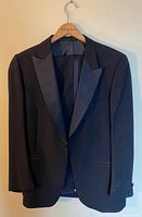 Black tuxedo jacket hanging on wooden hanger showing front and shoulders.