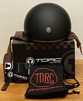 Front view of TORC T5 helmet in flat black, showing smooth rounded shell, padded brown chin strap with D-ring, resting on TORC box with carry bag and paperwork.