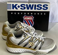 Front angle side view of a pair of white, black, and gold K-Swiss Big Shot women's tennis shoes with original black K-Swiss Performance box behind.