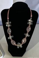 Full view of pink glass bead necklace displayed on black stand, showing faceted oval pink beads, clear cluster beads, and pink cord.