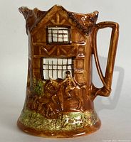 Front side view of brown ceramic pitcher shaped like a cottage with raised hunting scene detail showing a man on horseback and dogs in front of a timbered cottage.