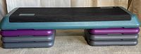 Aerobic step platform on six risers, viewed frontally on a carpeted floor against a white backdrop.