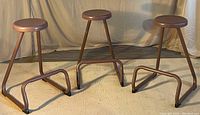 Photo showing three brown metal stools arranged side by side on a striped carpet with beige backdrop, displaying trapezoidal tubular bases and round seats.