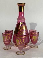 Photo showing entire set of a pink crystal decanter and six matching glasses, all decorated with gold leaf floral designs.