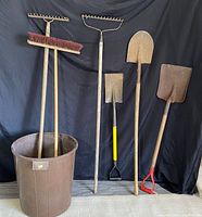 Full lot view showing two rakes, push broom, three shovels and garbage bin against black backdrop