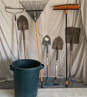 Full lot of two metal rakes, four metal shovels, one heavy-duty push broom, and one green plastic garbage bin