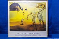 Full view of framed Salvador Dali Museum poster with surrealist yellow and brown painting titled 'Enchanted Beach with Three Fluid Graces' showing three stylized female figures on a light blue background.