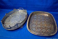 Photo showing two antique silver plated serving trays with tarnish. One is an oval footed tray with pierced sides and handles. The other is a rectangular etched tray with a patterned surface.