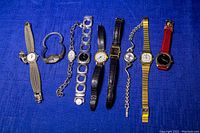 Full view of 9 assorted vintage women's wristwatches on a blue fabric background with various band and dial styles including brands Movado and Terner