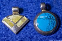 Photo showing two sterling silver pendants on a blue fabric background: round turquoise pendant and fan-shaped opalescent pendant.