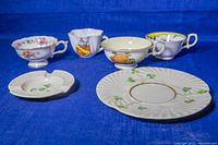 Photo of five teacups, one saucer, and one ashtray with varied floral and fruit designs and shamrock embossed porcelain pieces