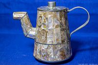 Metal teapot with handle and spout, showing abalone shell inlay details on the body and lid, photographed against a blue background.