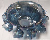 Blue iridescent carnival glass punch bowl with scalloped rim and attached hooks holding ten matching cups, showing the set as a whole.