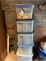 Stack of five large plastic storage bins with various lids, some clear with purple lids, others opaque with black lids. Bins contain wrapped home decor items and household goods.