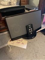 Bose SoundDock speaker with black mesh grille on front, sitting on charging dock or base, accompanied by Bose remote control placed on dock.