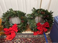 Two artificial Christmas wreaths displayed against a white door on a carpet with traditional patterns. Both have pine cones, red berries, and red bows, with string lights around them.
