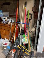 Photo showing assortment of garden and lawn tools including rakes, shovels, broom, sprayer, and others grouped and leaning against a wall in a garage area.