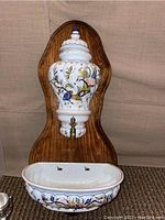 Hand painted French faience pottery mounted on a wood backing with a walnut finish, featuring floral and butterfly designs and a small basin beneath with two holes for drainage or mounting.