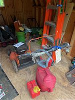 Husqvarna orange rotary lawn mower in shed with owner's manual on lawn mower deck, wrapped handlebar, and two red gas containers on floor in front