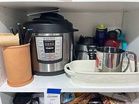 Shelving with Instant Pot Plus multi-cooker, utensil holder with wooden mallets and pens, Oster blender base, white ceramic dish, stainless steel sifter, and plastic water bottles