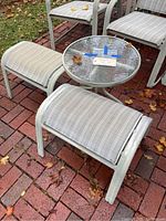Photo showing two outdoor stools with tubular metal frames and light striped fabric seats beside a round glass top end table with a light-colored metal frame