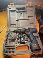 Open gray molded plastic case showing Ryobi 6.0V cordless drill, charger, and assorted drill bits.