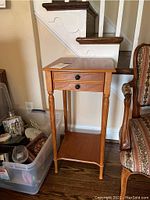 Full view of the tall side table showing the maple stain finish, drawers, and lower shelf next to stair railing and upholstered chair.