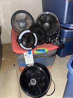 Photo showing three tabletop fans and two clip-on mini fans placed on a large plastic container in basement storage. The fans vary in size and design, primarily in black color with one Holmes branded fan visible.