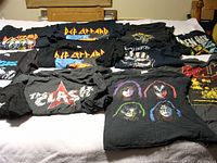 Eleven black rock band T-shirts laid out on a bed featuring Def Leppard, Kiss, Guns N' Roses, The Clash, and others with band logos and artwork.