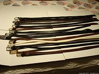 Full view of 14 men's belts laid out flat showing various colors, lengths, and buckle styles on white cardboard background.