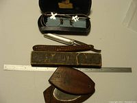 Top down photo of spectacles in case above straight razor, strop and wooden mirror close to ruler for scale