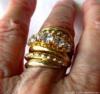 Close-up photo of the gold-gilded sterling silver ring on finger, showing five round clear cubic zirconia stones prong set in a wide, decorated band with openwork design, wire brushed finish, and hammered elements.